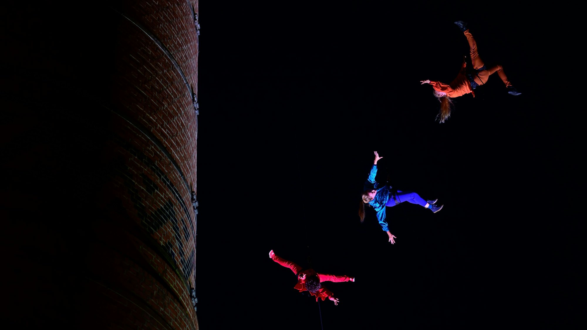 Drei Personen schwingen an Seilen um einen Backsteinturm.