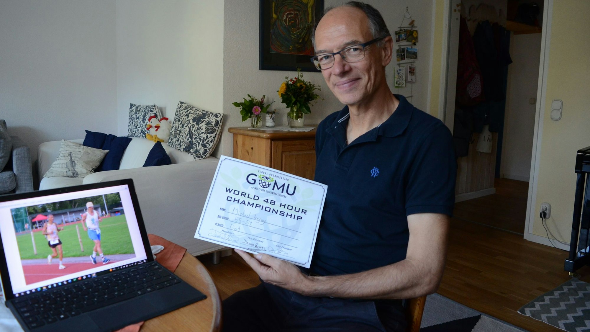 Der Mann sitzt an einem Tisch in einem Wohnzimmer und hält eine Urkunde in die Höhe.