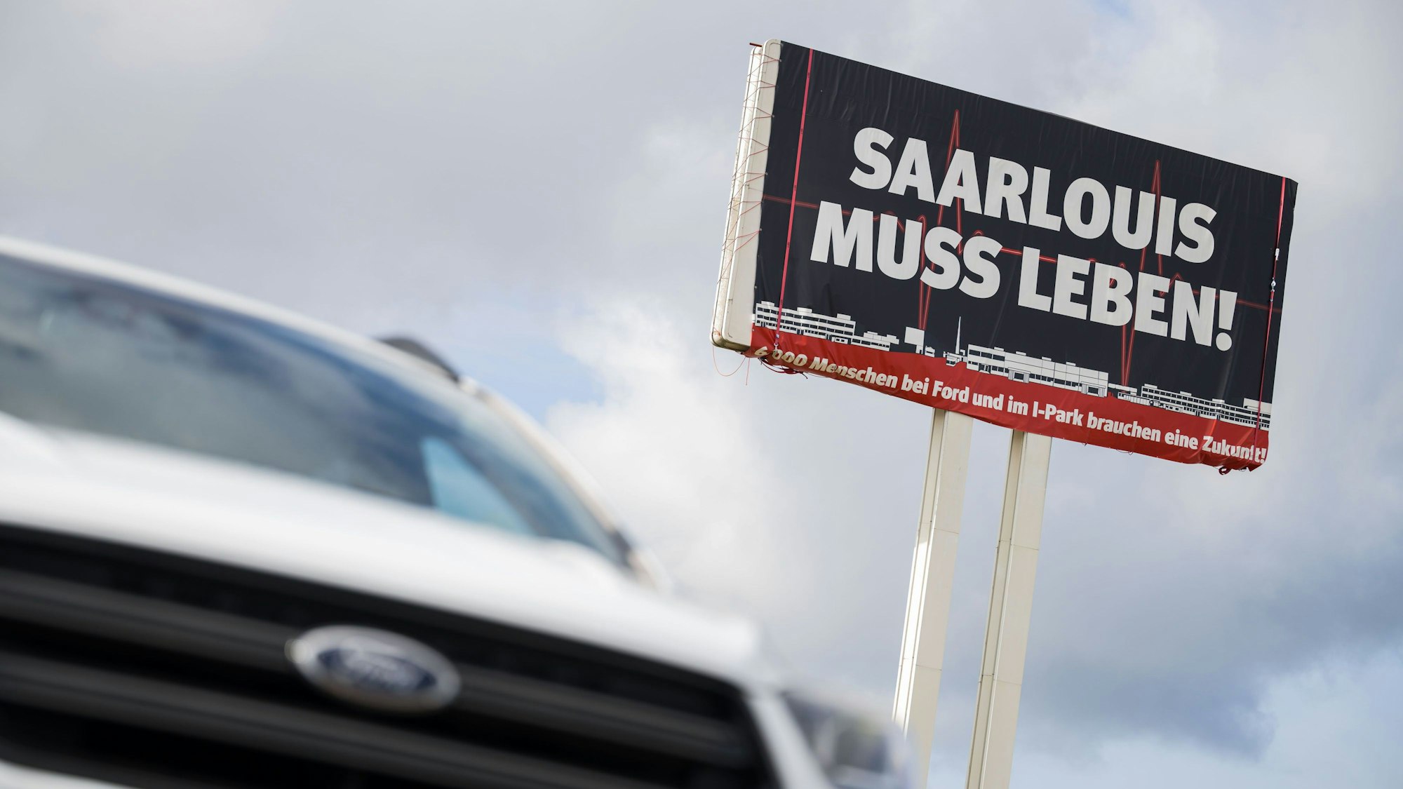 Ein Ford Kuga steht vor einem großen Schild auf dem für die Bedeutung des Ford Werks in Saarlouis geworben wird.