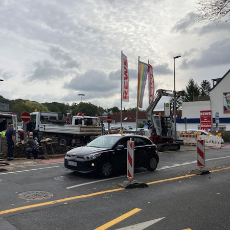 Zu sehen sind Baufahrzeuge und Absperrbaken vor dem Supermarktparkplatz. Daran vorbei fährt ein Kleinwagen.