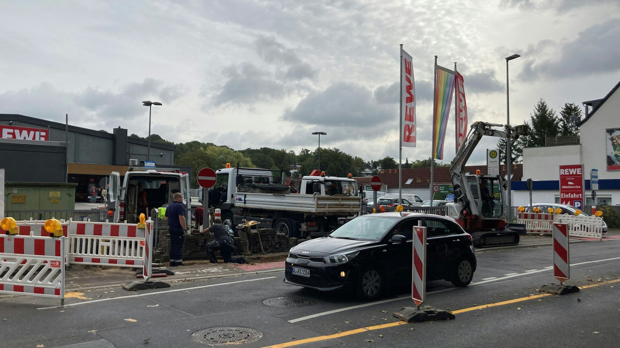 Zu sehen sind Baufahrzeuge und Absperrbaken vor dem Supermarktparkplatz. Daran vorbei fährt ein Kleinwagen.