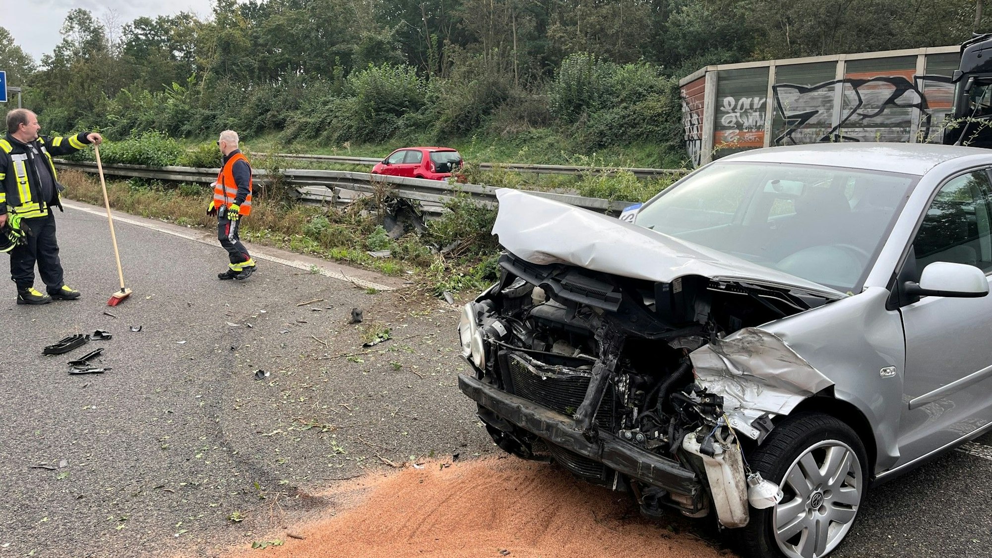 Ein silberner Pkw steht mit dem Heck an der eingedellten Mittelleitplanke, die Front ist verbogen, der Kühlergrill abgerissen. Ausgelaufene Betriebsstoffe wurden mit rotem Bindemittel abgestreut. Zwei Männer mit Warnwesten und Besen stehen auf der Fahrbahn.