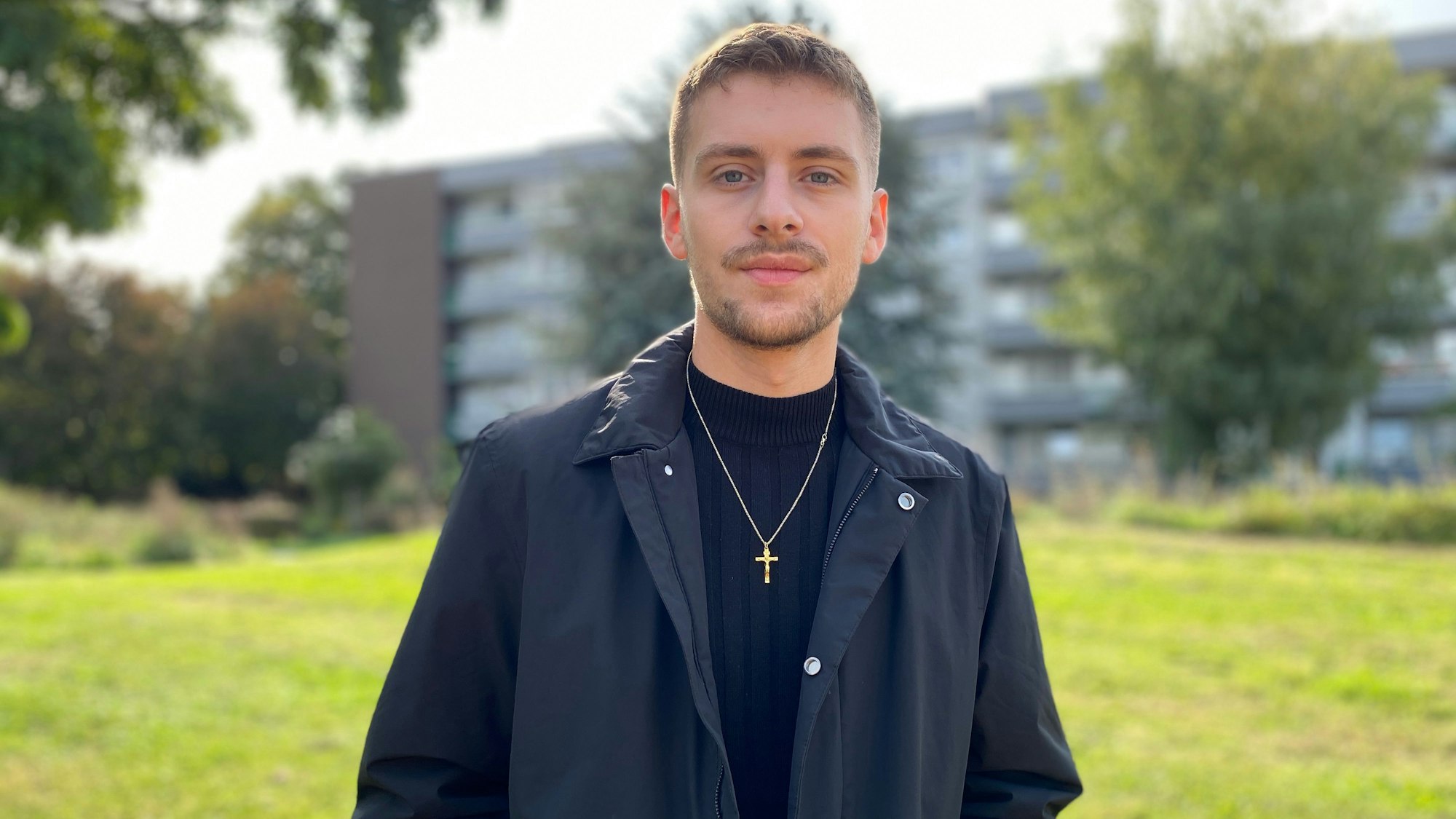 Ein junger Mann in dunkler Kleidung mit Dreitagebart und Kreuz-Anhänger auf einer Wiese vor einer Wohnsiedlung.