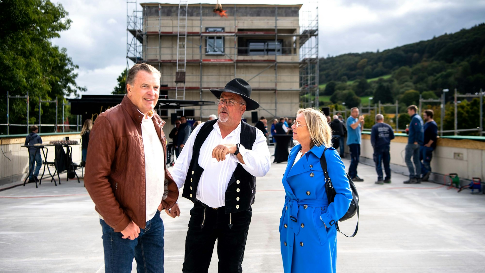 Bauherr Marc Brucherseifer (l.), Zimmermeister Jürgen Dreßen und Bürgermeisterin Sabine Preiser-Marian stehen auf einem betonierten Platz vor einem eingerüsteten und in Bau befindlichen Gebäude.