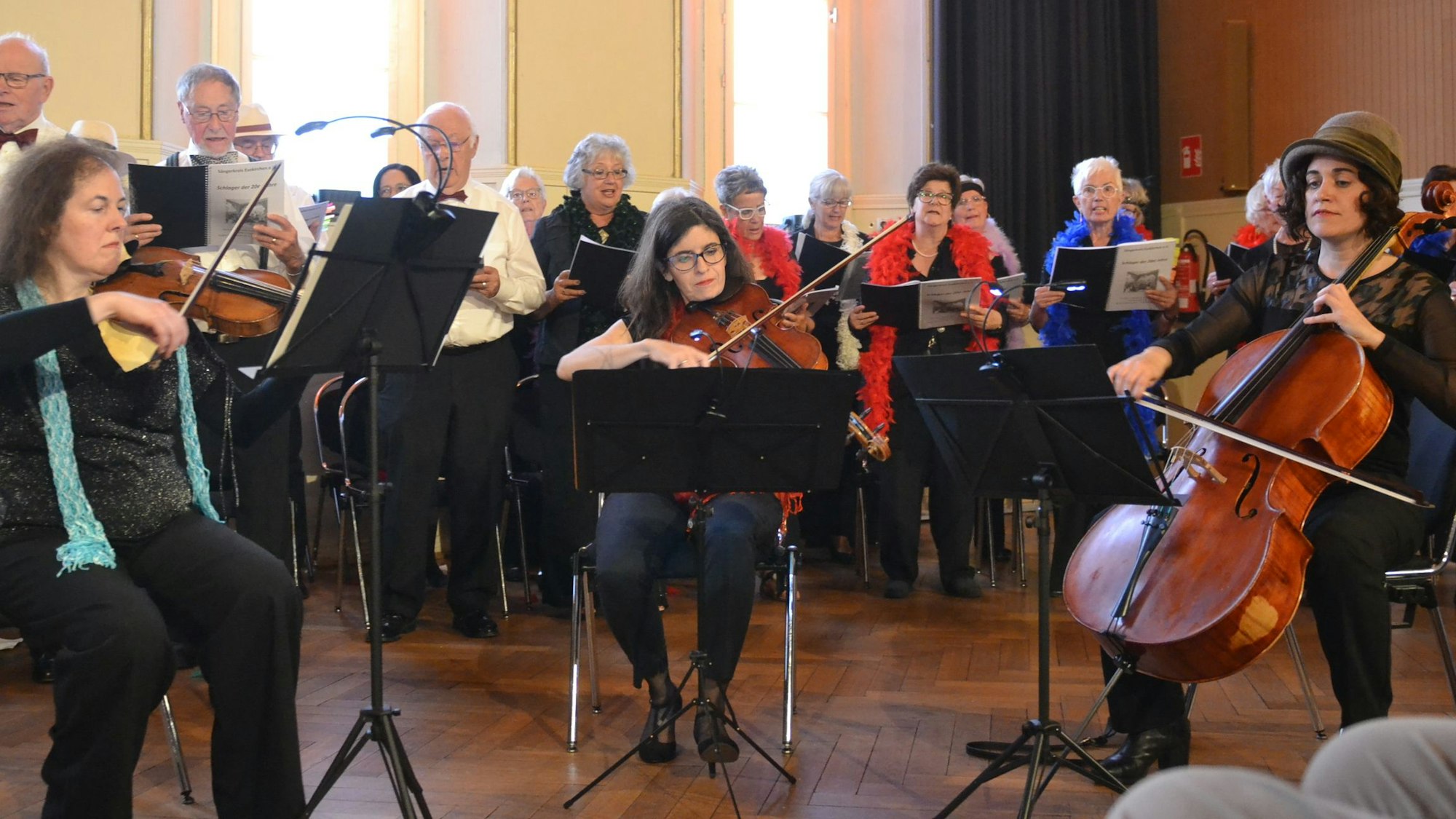 Im Vordergrund musizieren die drei Frauen des Salon-Orchesters, im Hintergrund singt der Chor.