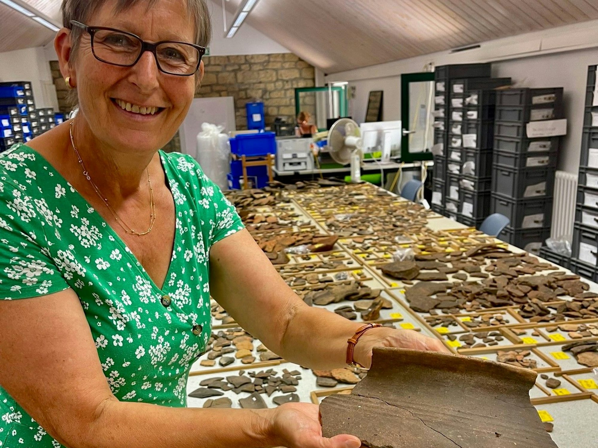 Eine Frau zeigt Tonscherben, weitere sind auch einem langen Tisch ausgebreitet.