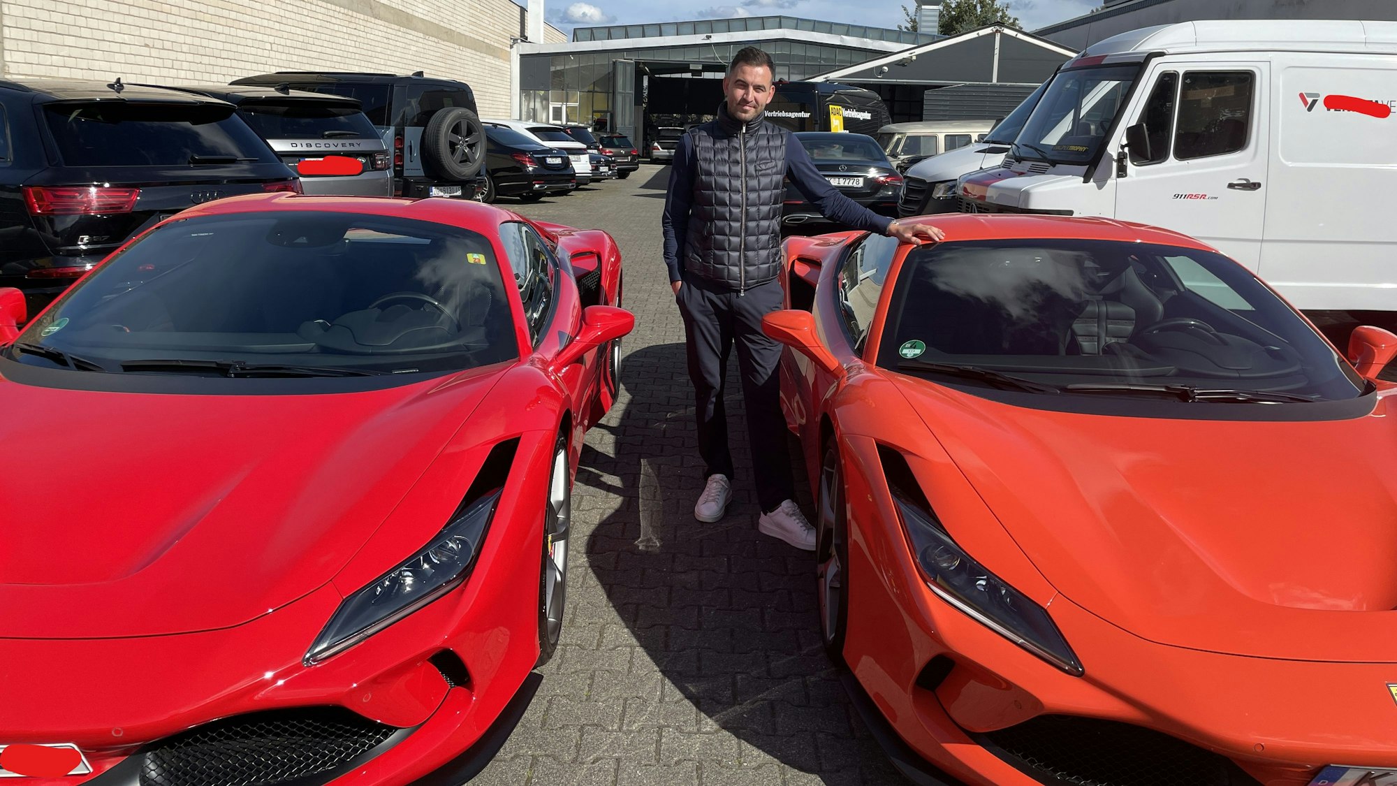 Ein junger Mann mit dunklem Haar steht zwischen zwei roten Ferrari.