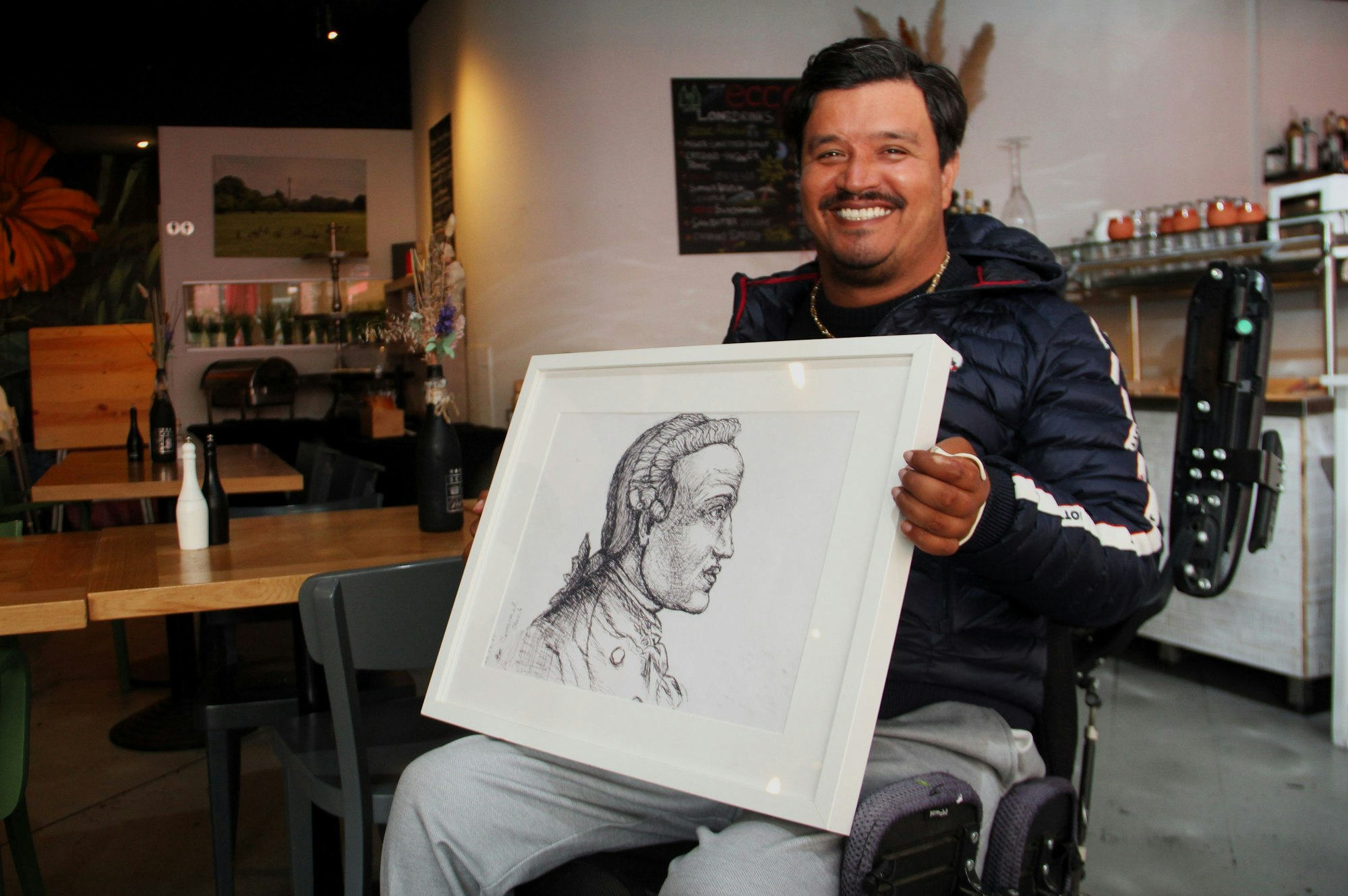 Ein dunkelhaariger sitzt in einem Rollstuhl, auf dem Schoß hält er ein Schwarz-Weiß-Porträt.