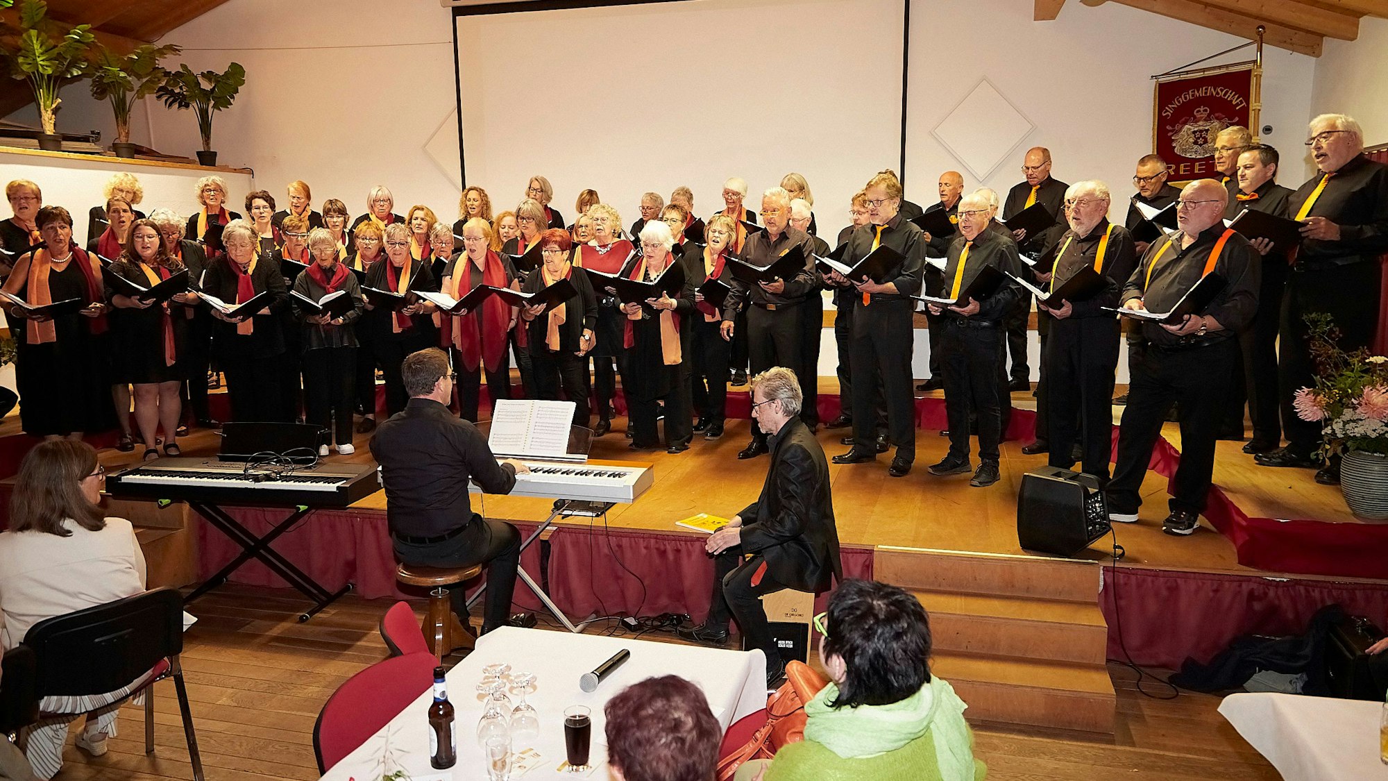 Sängerinnen und Sänger eines Chores stehen auf einer Bühne und singen. Davor sind einige Tische des Saals zu erkennen.