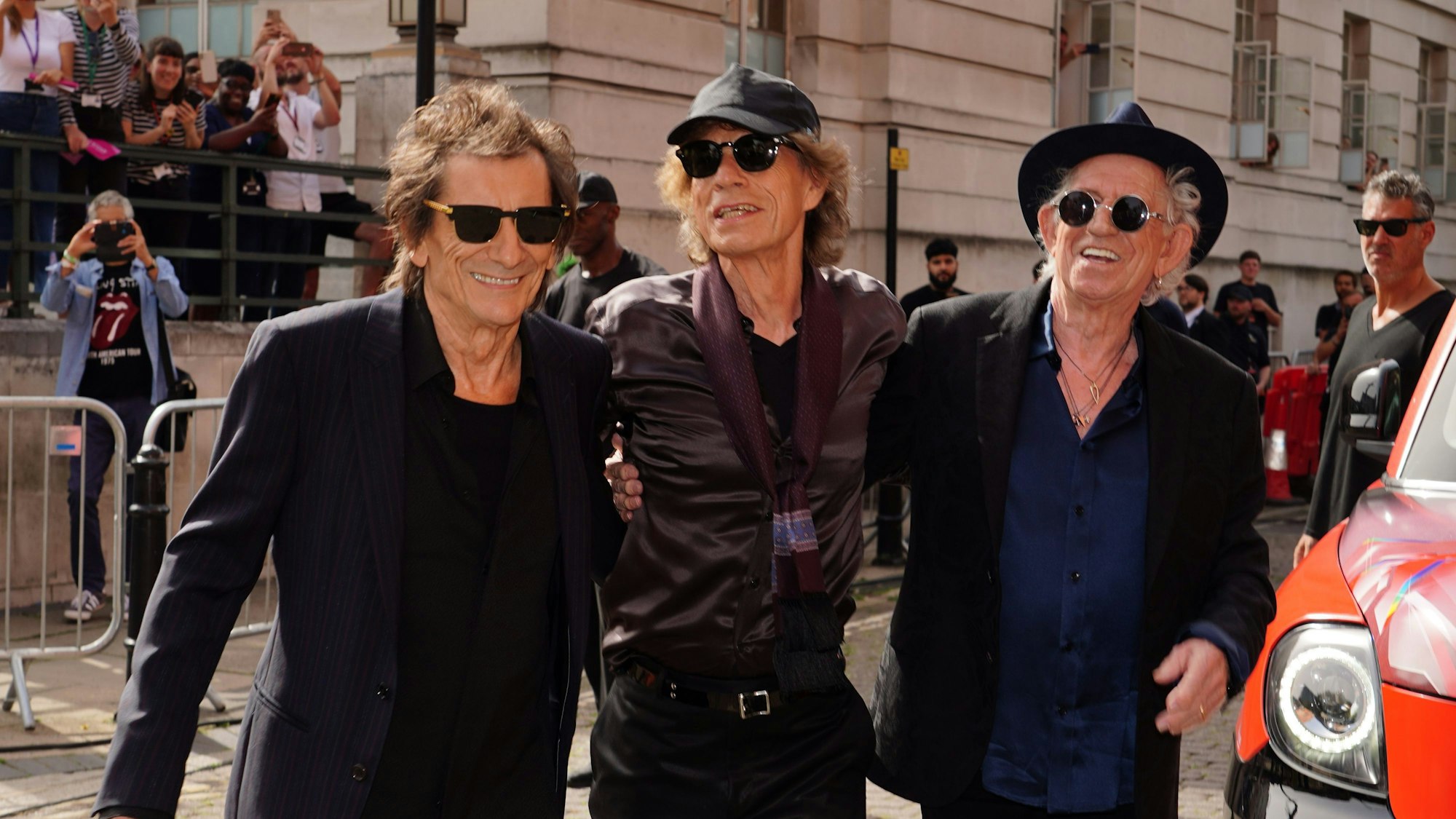 Die drei Bandmitglieder stehen Arm in Arm. Im Hintergrund sind Fans und Fotografen zu sehen.