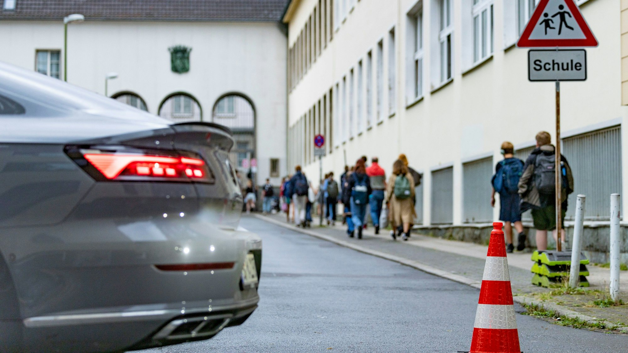 Vor einer Schule stehen Pylonen auf der Straße. Schüler und Schülerinnen gehen zu Fuß über den Gehweg.
