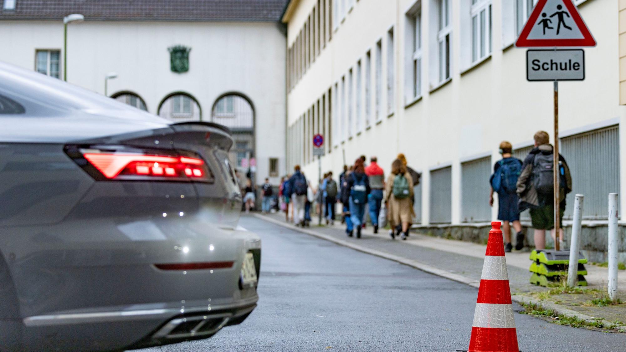Vor einer Schule stehen Pylonen auf der Straße. Schüler und Schülerinnen gehen zu Fuß über den Gehweg.