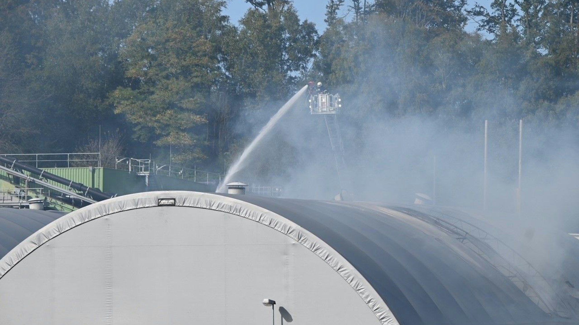 Ein Feuerwehrmann löscht von einer Drehleiter aus eine brennende Halle mit Zeltdach.