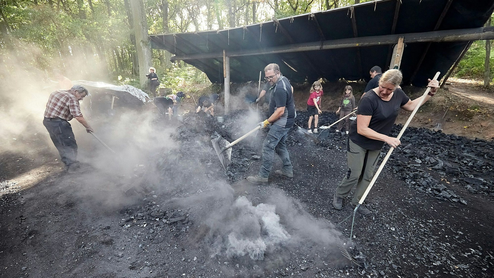 Frauen, Männer und Kinder arbeiten an einem Kohlenmeiler, um sie herum ist Rauch.