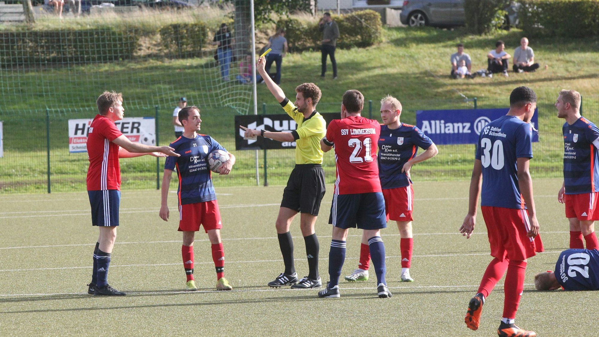 Der Schiedsrichter zeigt einem Fußballer die gelbe Karte. Ein Mitspieler protestiert.Ein gegnerischer Spieler liegt auf dem Boden.