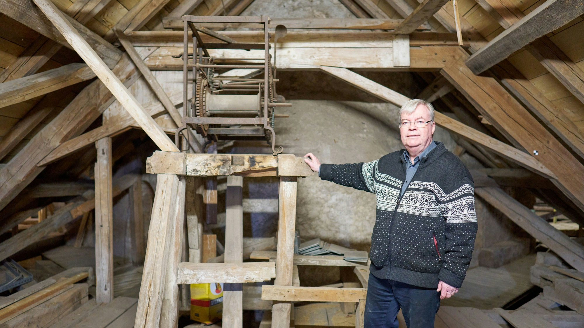 Helmut J. Kirfel steht vor einem Holzgerüst, auf der die Reste einer Uhr aus dem 15. Jahrhundert auf dem Dachboden der Steinfelder Basilika zu sehen sind.