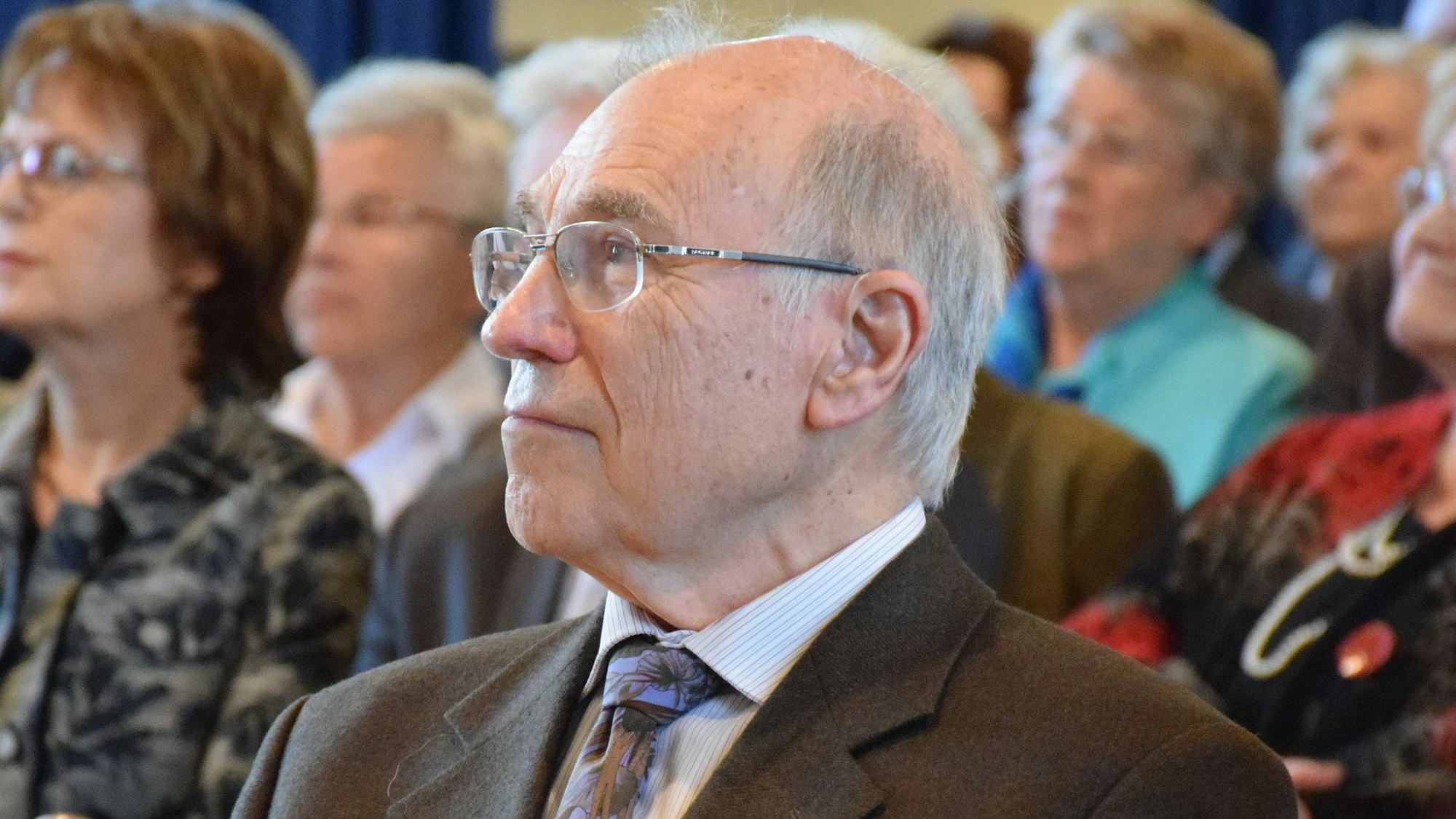 Archivfoto von Wolfram Königsfeld. Er trägt eine Brille und einen Anzug, im Hintergrund sind weitere Personen erkennbar.