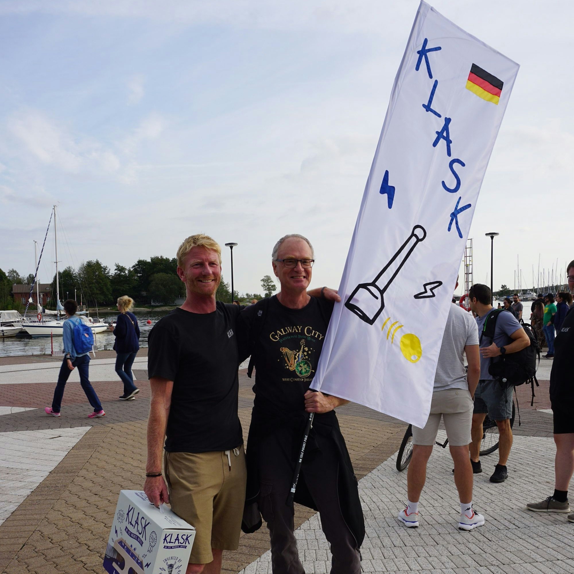 Klask-Erfinder Mikkel Bertelsen und Ingo Ebel stehen nebeneinander. Ebel hält eine Fahne in der Hand, auf der „Klask“ steht und die Deutschland-Flagge gemalt ist.