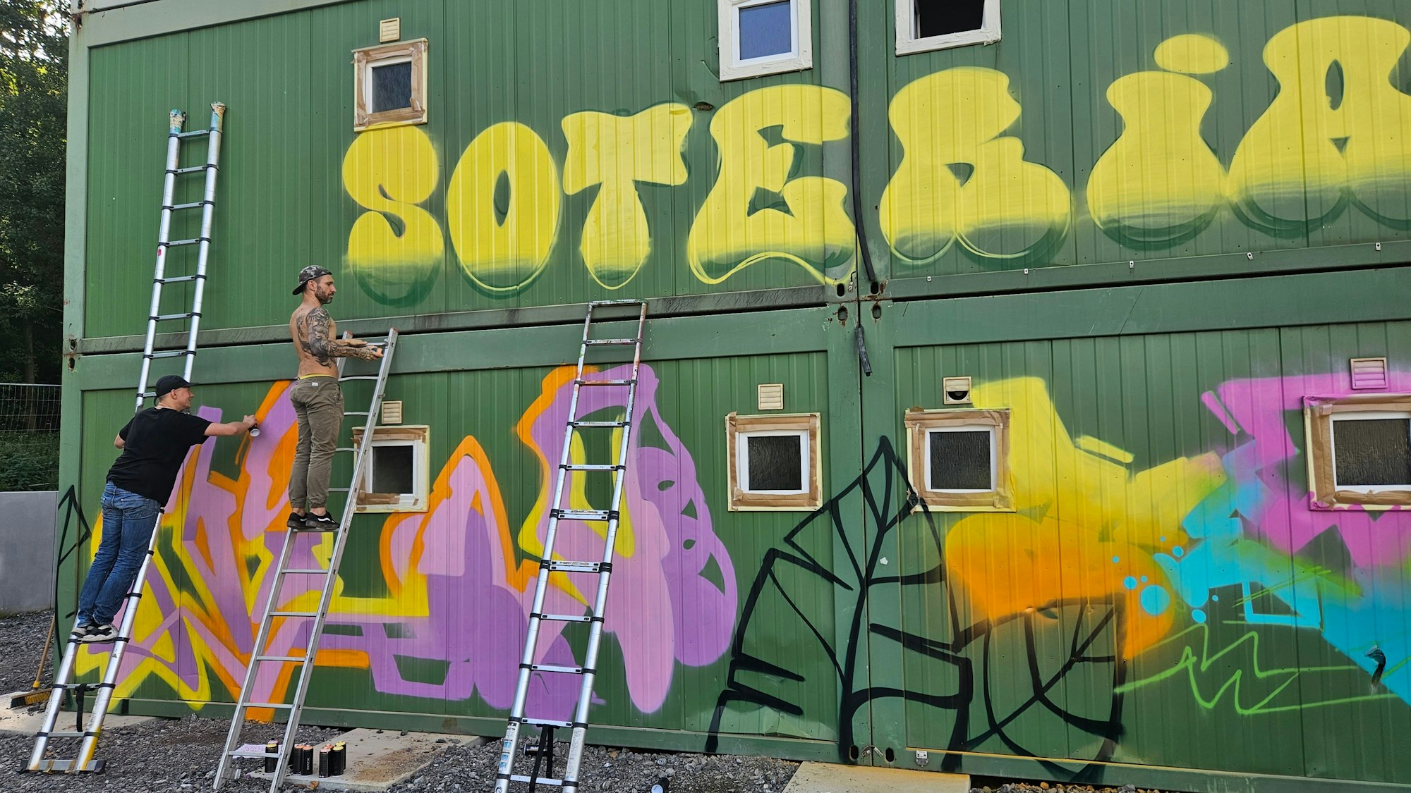 Zwei Graffiti-Künstler stehen auf Leitern und besprühen Container der neuen Wohnanlage für Flutopfer in Gemünd.