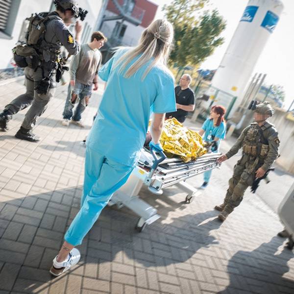 Zwei Mitglieder einer Spezialeinheit in voller Kampfmontur und zwei Mitarbeiterinnen der Notaufnahme schieben eine Krankentransportliege mit dem Darsteller eines Verletzten, der unter einer goldenen Rettungsfolie liegt.