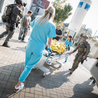 Zwei Mitglieder einer Spezialeinheit in voller Kampfmontur und zwei Mitarbeiterinnen der Notaufnahme schieben eine Krankentransportliege mit dem Darsteller eines Verletzten, der unter einer goldenen Rettungsfolie liegt.