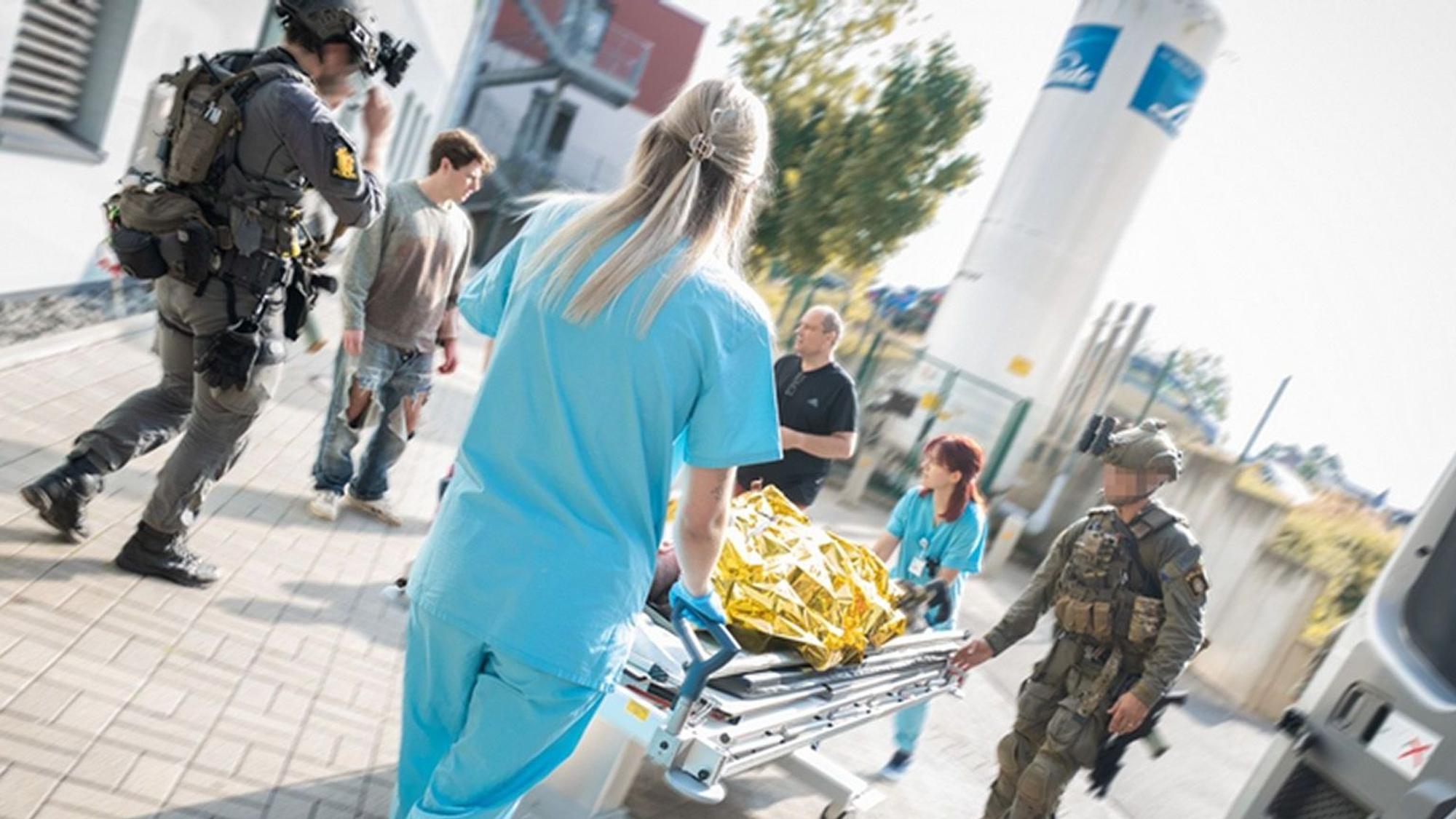 Zwei Mitglieder einer Spezialeinheit in voller Kampfmontur und zwei Mitarbeiterinnen der Notaufnahme schieben eine Krankentransportliege mit dem Darsteller eines Verletzten, der unter einer goldenen Rettungsfolie liegt.