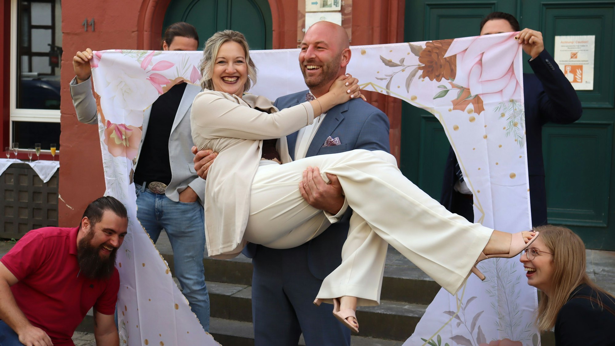 Tim Schmitt trägt seine frisch angetraute Frau Jenny Ulhaas. Im Hintergrund ein Leinentuch mit einer herzförmigen Öffnung.