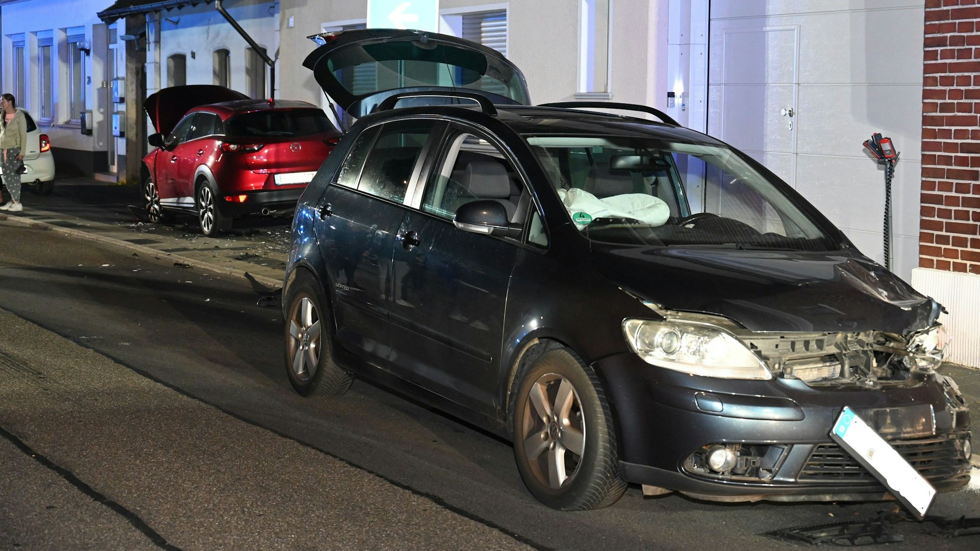 Ein schwarzes Auto steht an der Hauswand, die linke Seite eingedrückt. Im Hintergrund steht eines der beschädigten Fahrzeuge.