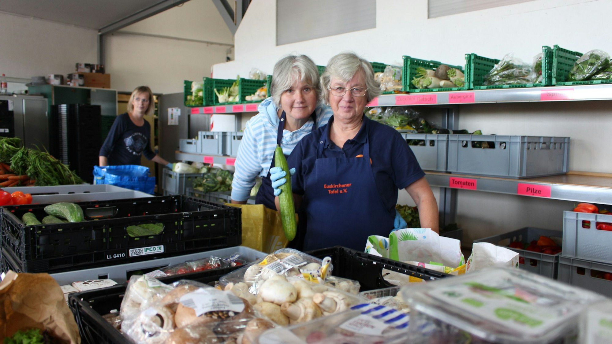 Drei Mitarbeiterinnen der Tafel laufen an den Kisten mit Obst und Gemüse vorbei, um die Taschen der Kunden zu füllen.