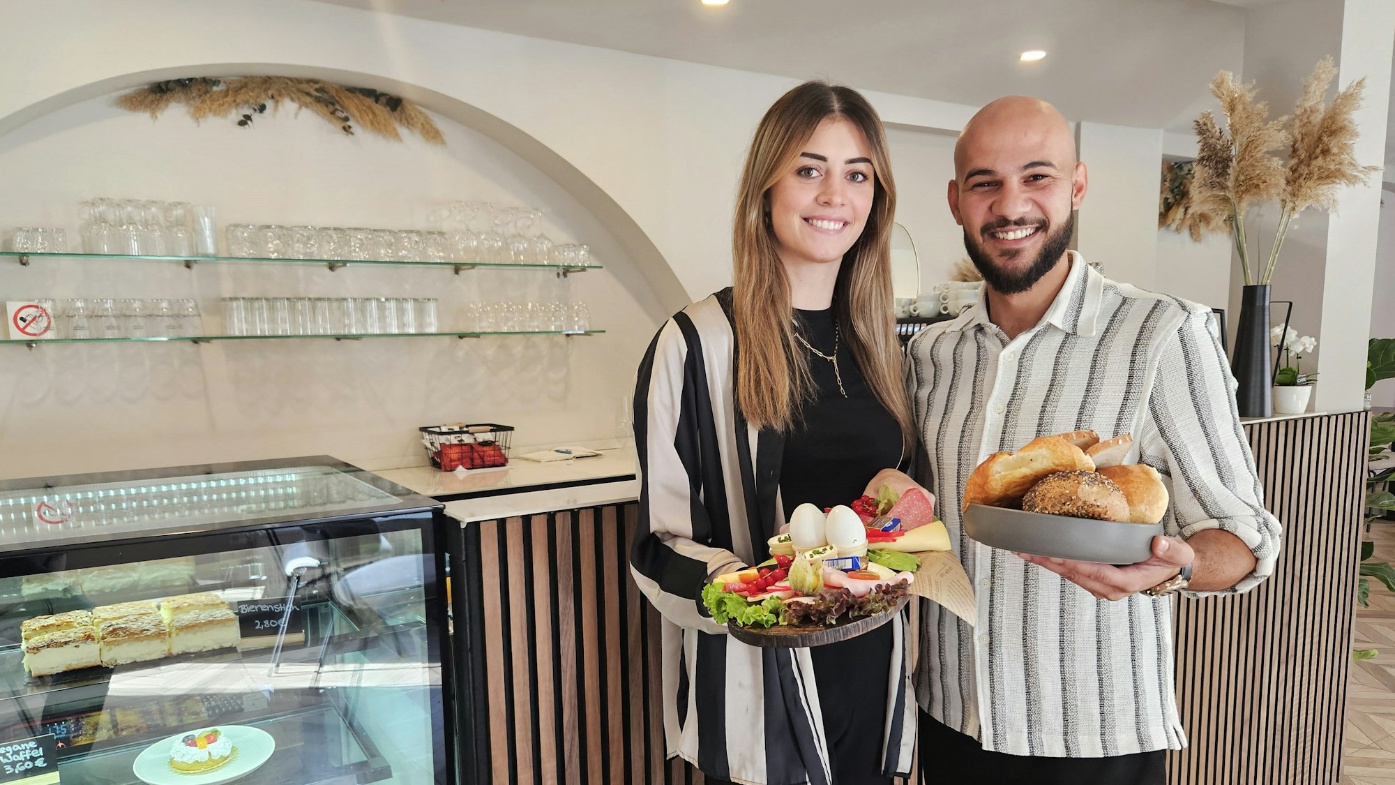 Linda und Ramadan Ajdini präsentieren vor der Theke ein Frühstück für zwei Personen.