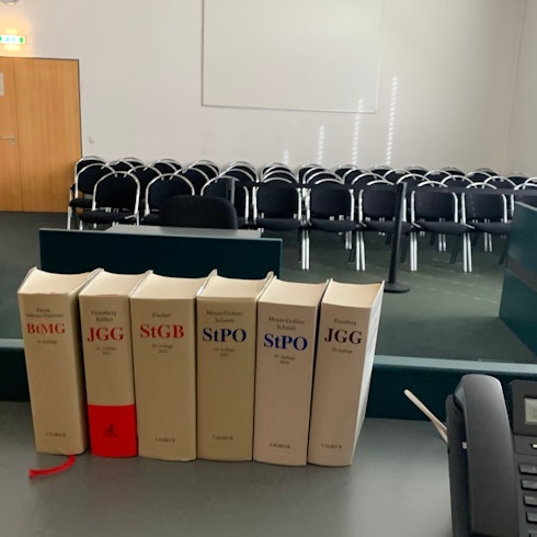 Das Symbolbild zeigt einen Gerichtssaal und im Vordergrund Bücher mit Gesetzestexten.