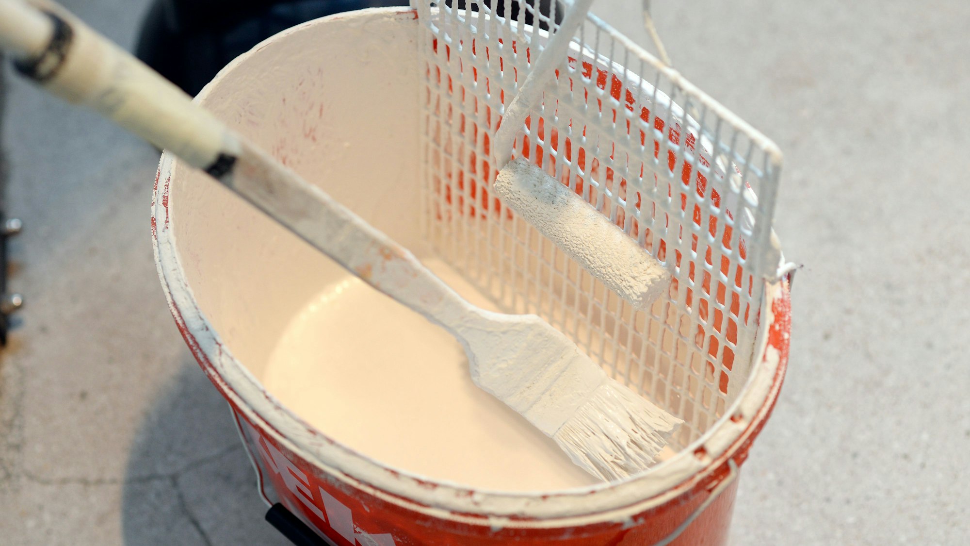 Das Symbolbild zeigt einen nahezu geleerten Eimer mit weißer Farbe. Darin stecken in Pinsel und ein Abstreifgitter.