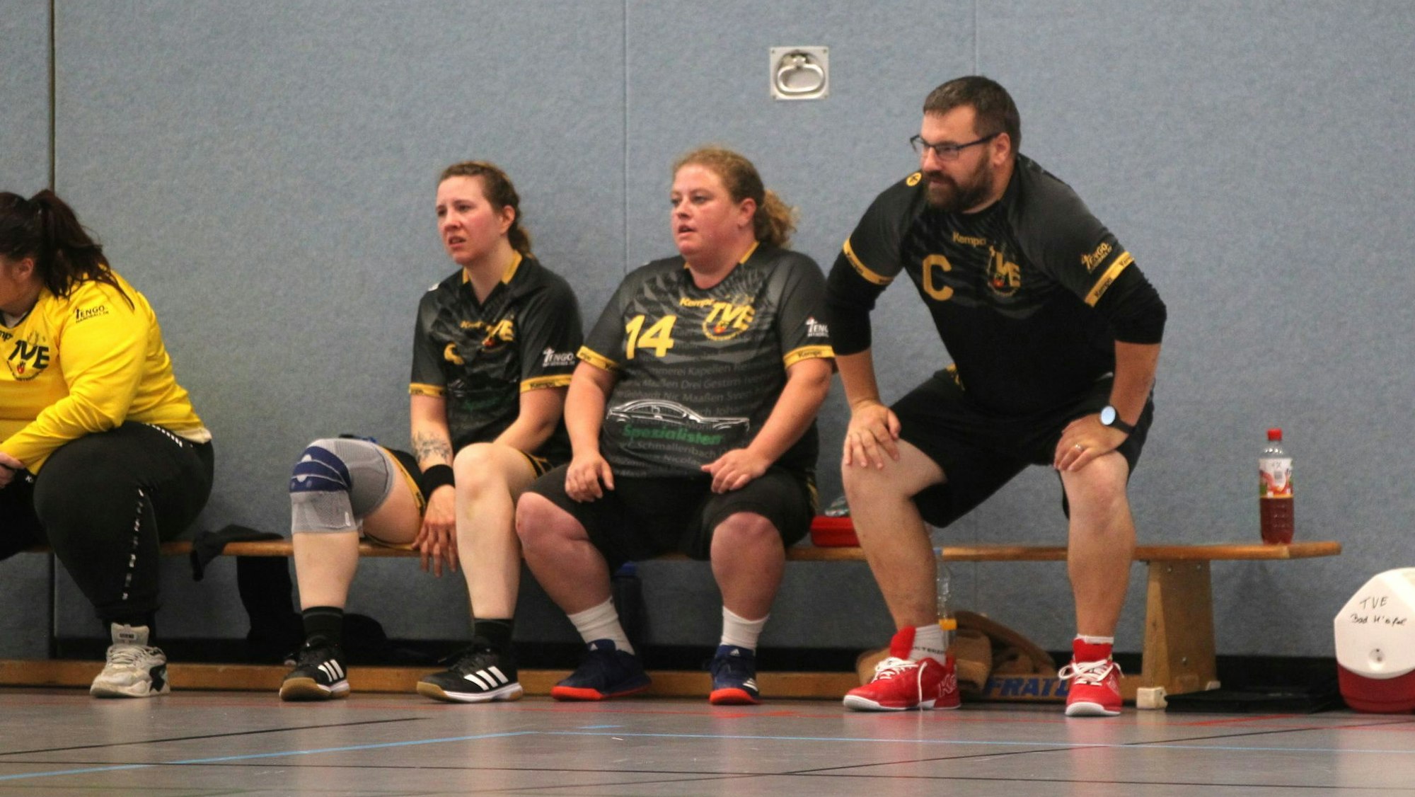 Die Auswechselbank einer Frauenhandballmannschaft. Der Trainer hockt davor, zwei Spielerinnen sitzen erschöpft auf der Bank..