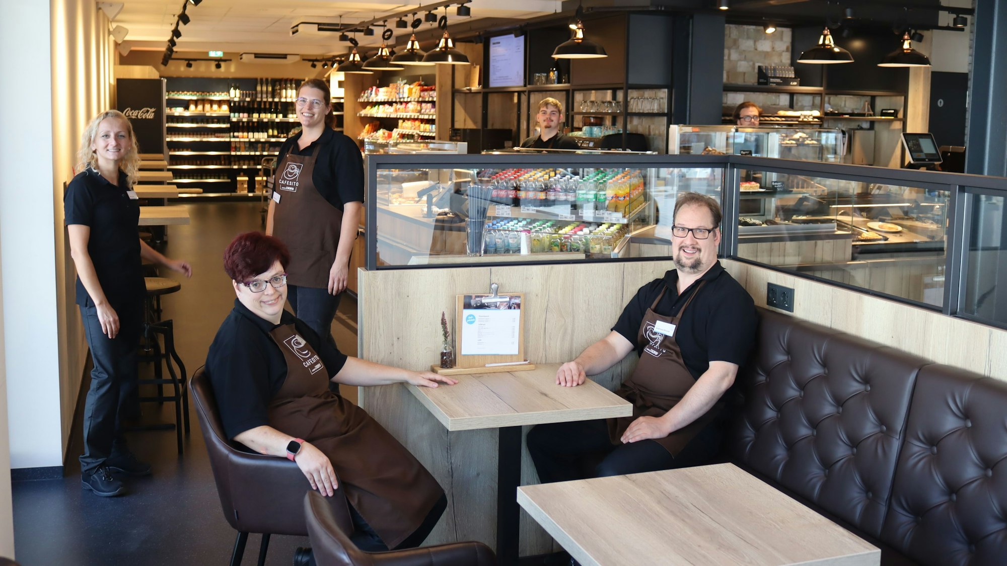 Die Nordeifelwerkstätten betreiben das Cafesito: Zwei Mitarbeiter sitzen an einem Tisch im Café, weitere stehen dahinter, im Hintergrund sind die Theke und der Verkaufsraum zu sehen.