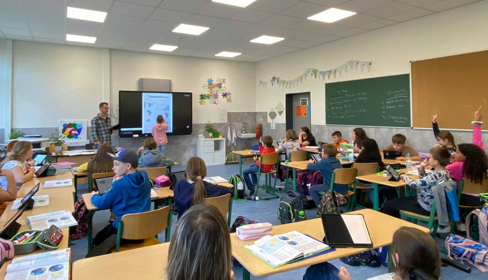 Ein Schülerin steht neben dem Lehrer an der digitalen Tafel. Die anderen sitzen auf ihren Plätzen und haben Tabletcomputer vor sich.