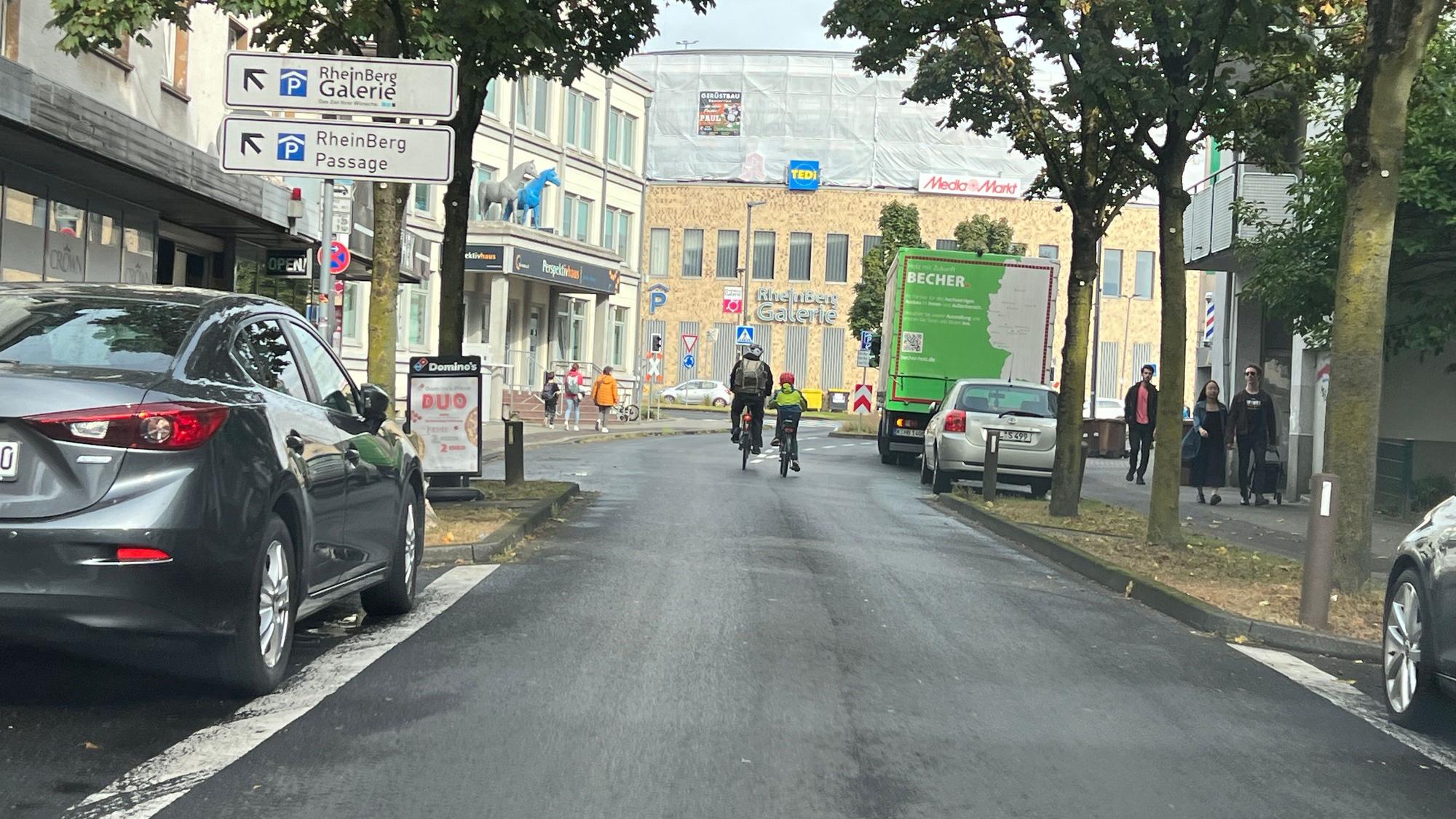 Zwei Radfahrer sind auf der unteren Hauptstraße in Richtung Schrankenkreisel und Rhein-Berg-Galerie unterwegs.