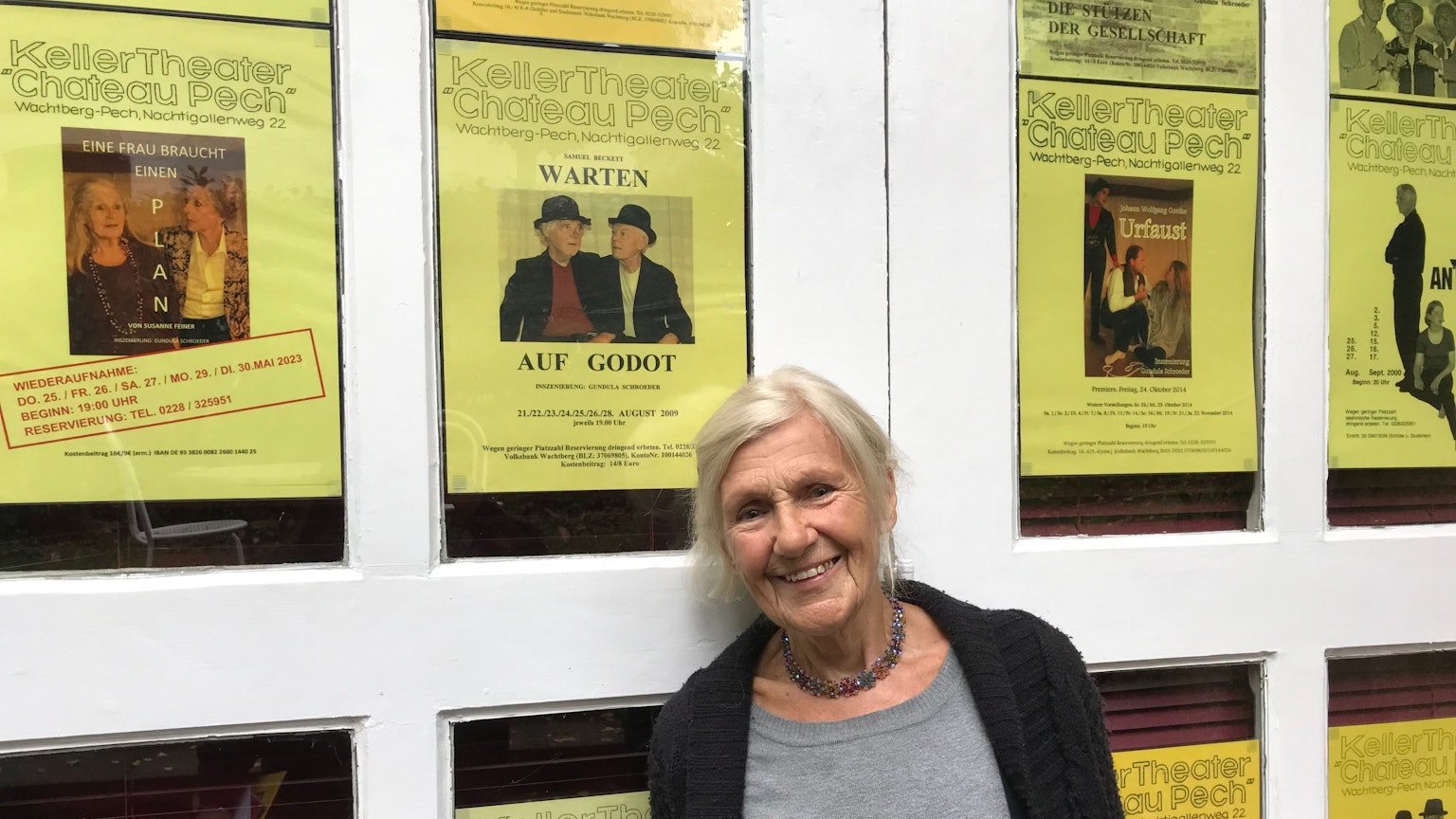 Gundula Schroeder steht vor einer Wand, an der Ankündigungsplakate einiger Aufführungen der vergangenen 25 Jahre hängen.