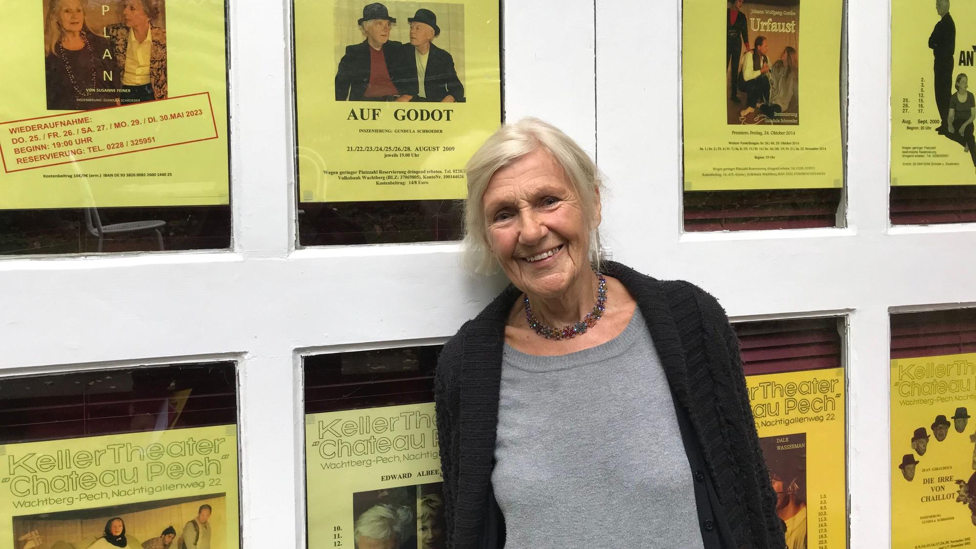 Gundula Schroeder steht vor einer Wand, an der Ankündigungsplakate einiger Aufführungen der vergangenen 25 Jahre hängen.