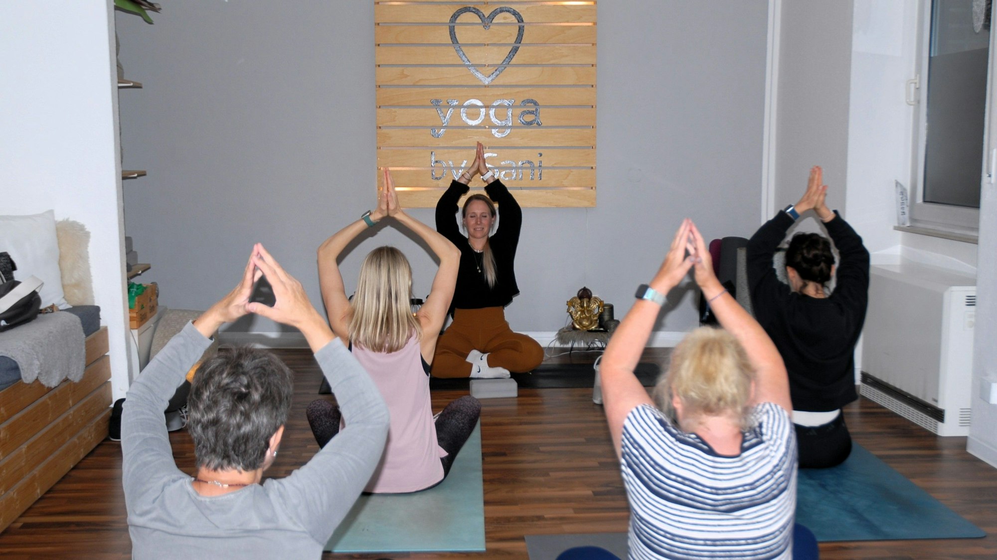 Frauen sitzen auf Matten im Schneidersitz und heben die Hände über den Kopf.