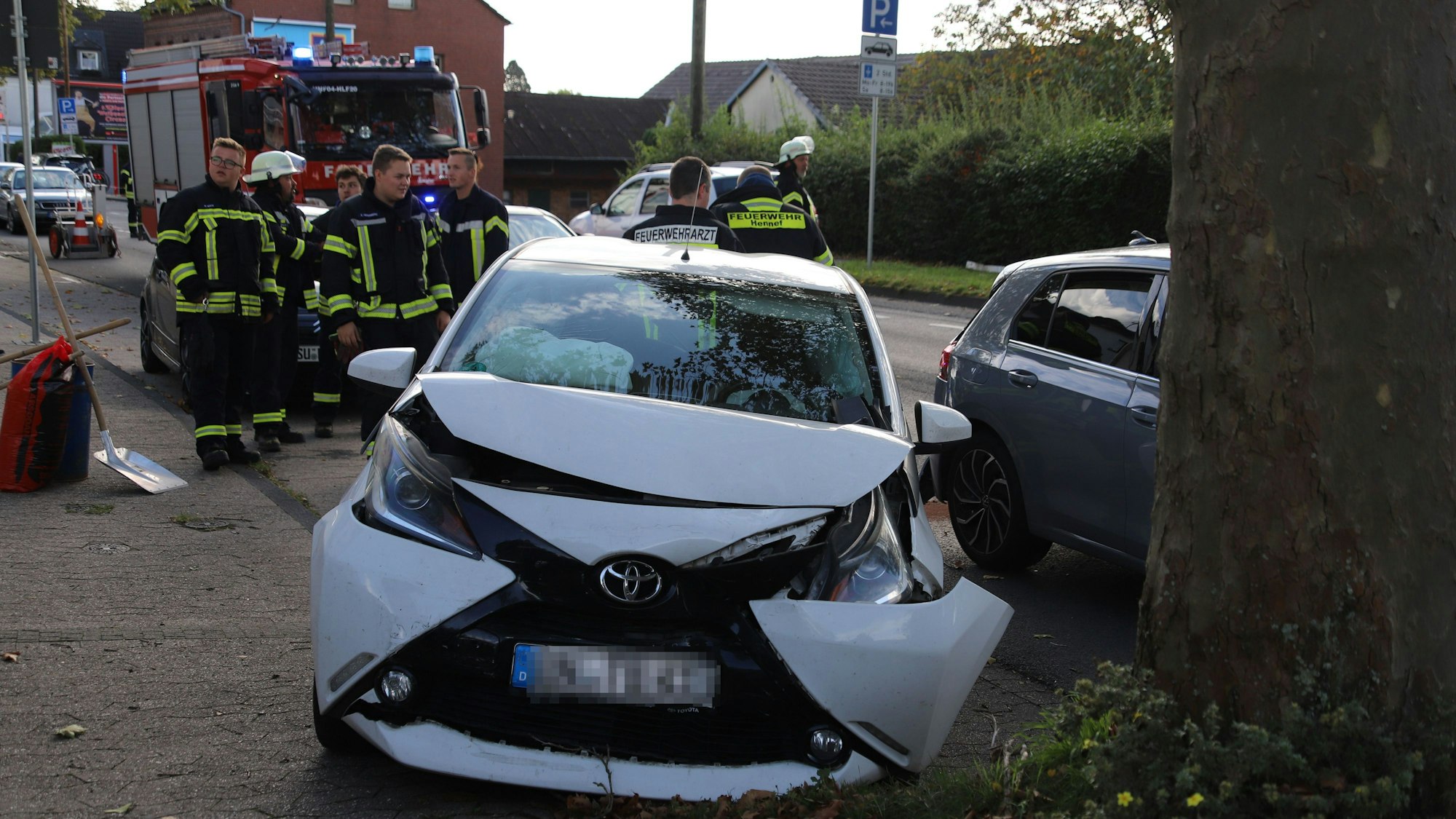 Ein Auto mit stark beschädigter Front steht neben einem Baum - im Hintergrund befinden sich Feuerwehrleute.