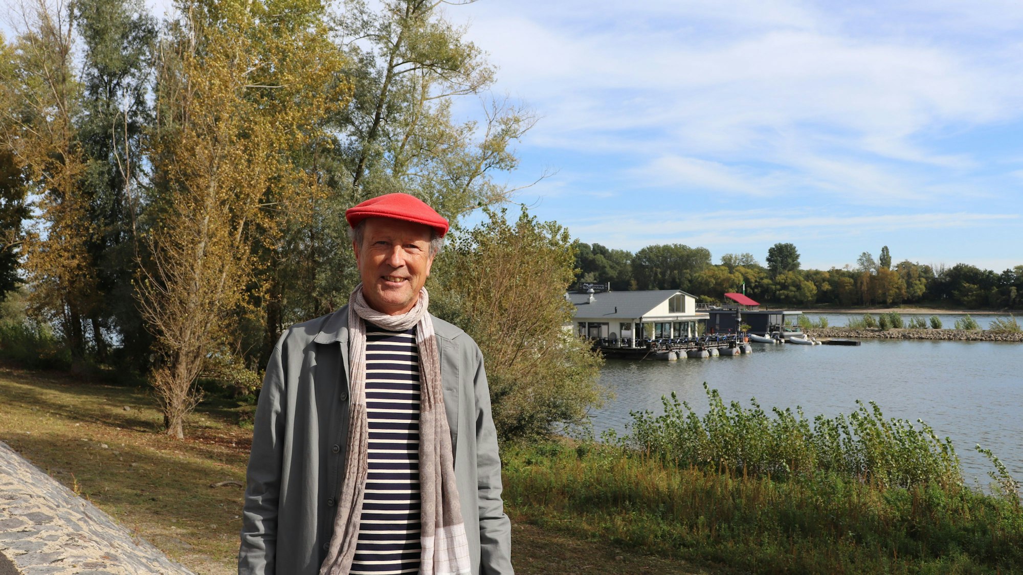 Ein Mann mit roter Baskenmütze auf dem Kopf steht am Rheinufer, im Hintergrund ist das Sürther Bootshaus zu sehen.