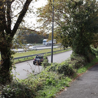 Ein Radweg liegt zwischen einer mehrspurigen Straße und einem umzäunten Gelände.