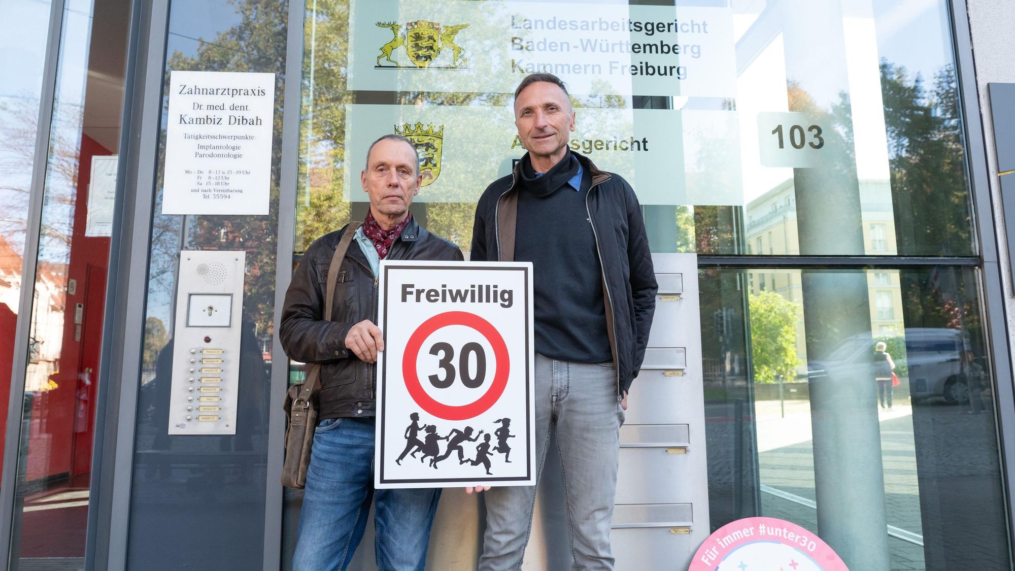 Christian Kronbitter (l) und Erich Maier halten vor dem Verwaltungsgericht ein Schild mit der Aufschrift „Freiwillig 30“ in die Kamera. Das Verwaltungsgericht Freiburg verhandelt über mehrere von der Deutschen Umwelthilfe (DUH) unterstützte Klagen zur Klärung der Frage, ob die Aufstellung von ·Freiwillig Tempo 30-Schildern rechtmäßig ist.