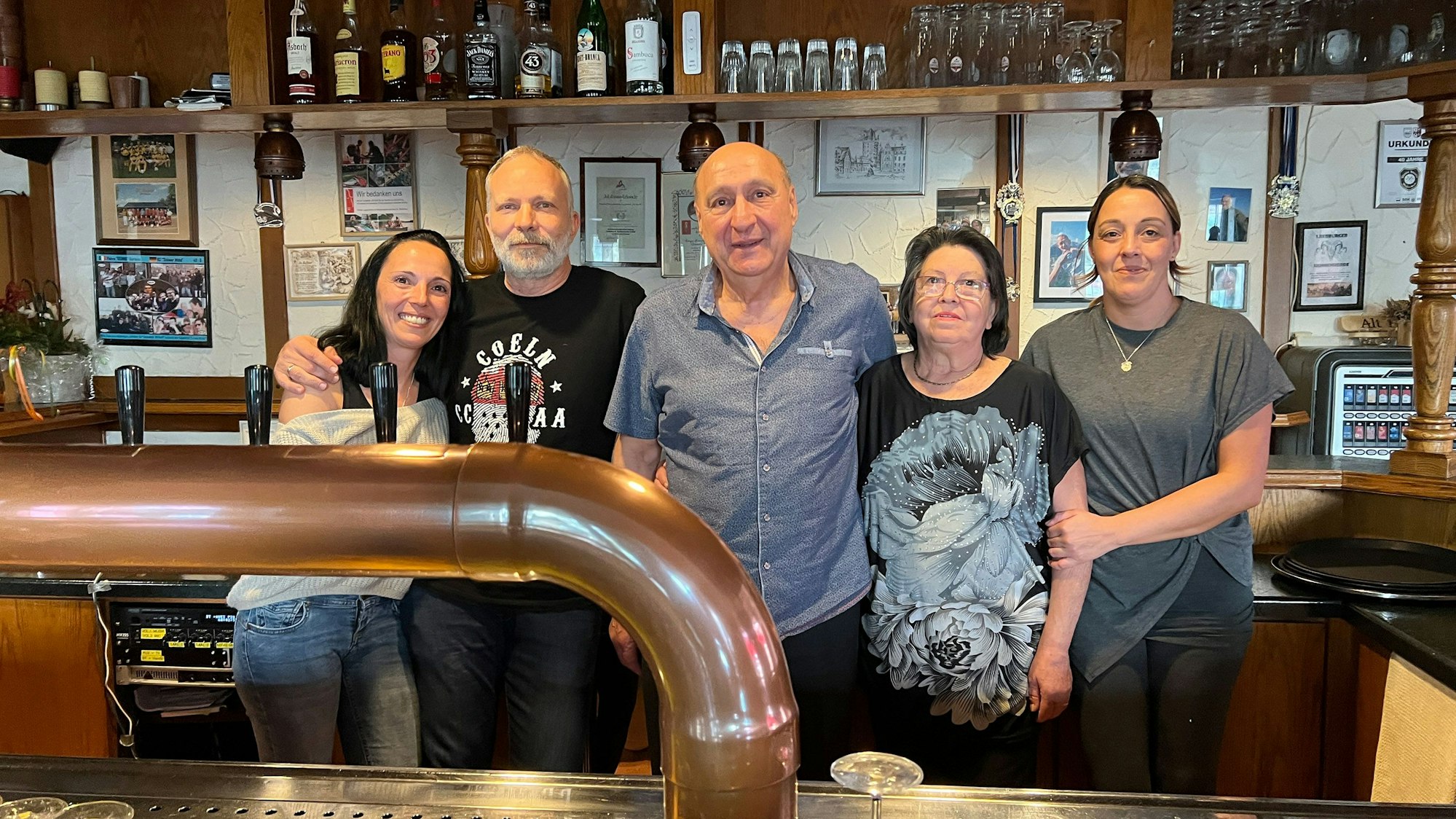 Zwei Männer und drei Frauen stehen hinter einer Theke, im Hintergrund stehen Gläser und Flaschen in einem offenen Schrank.
