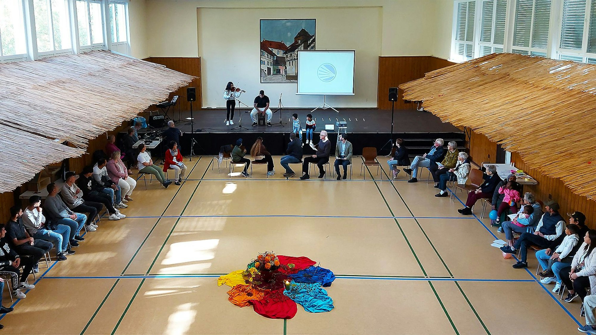 Zahlreiche Teilnehmende des Festes sitzen auf Stühlen in einem großen Kreis beisammen. Auf der Bühne treten zwei Musizierende auf.