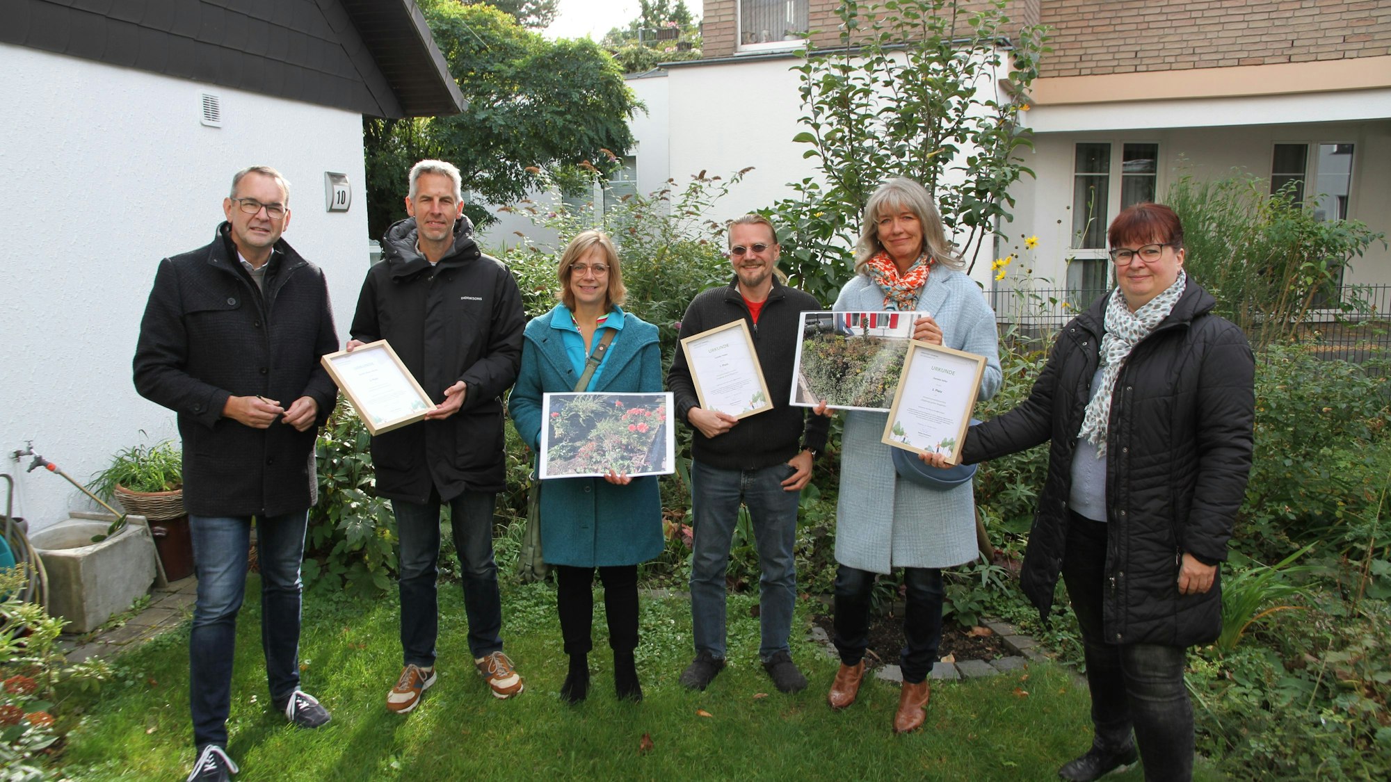 Drei Männer und drei Frauen stehen mit Fotos und Siegerurkunden in einem gepflegten Siegburger Garten