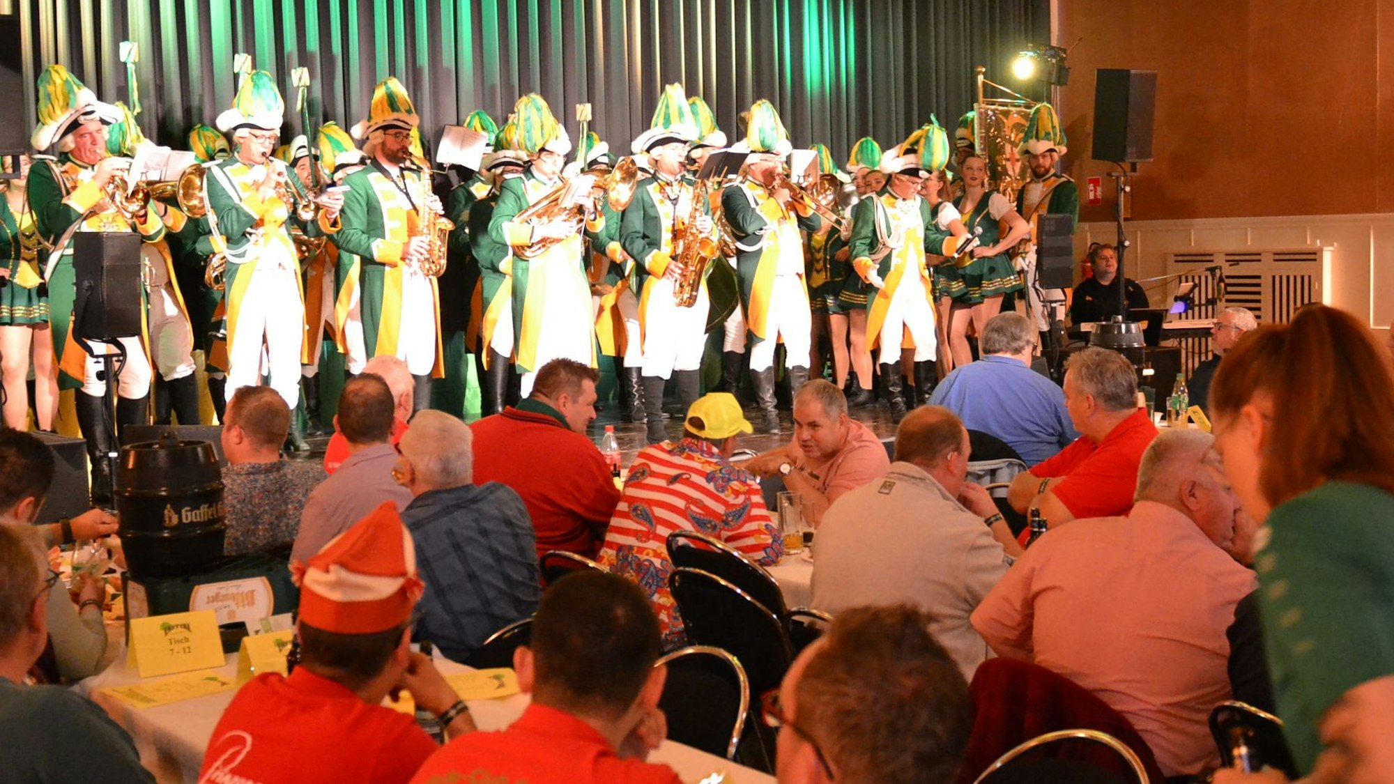 Auf der Bühne eines Veranstaltungssaals steht der Musikzug der Hovener Jungkarnevalisten. Davor sitzen zahlreiche Männer an Tischen.