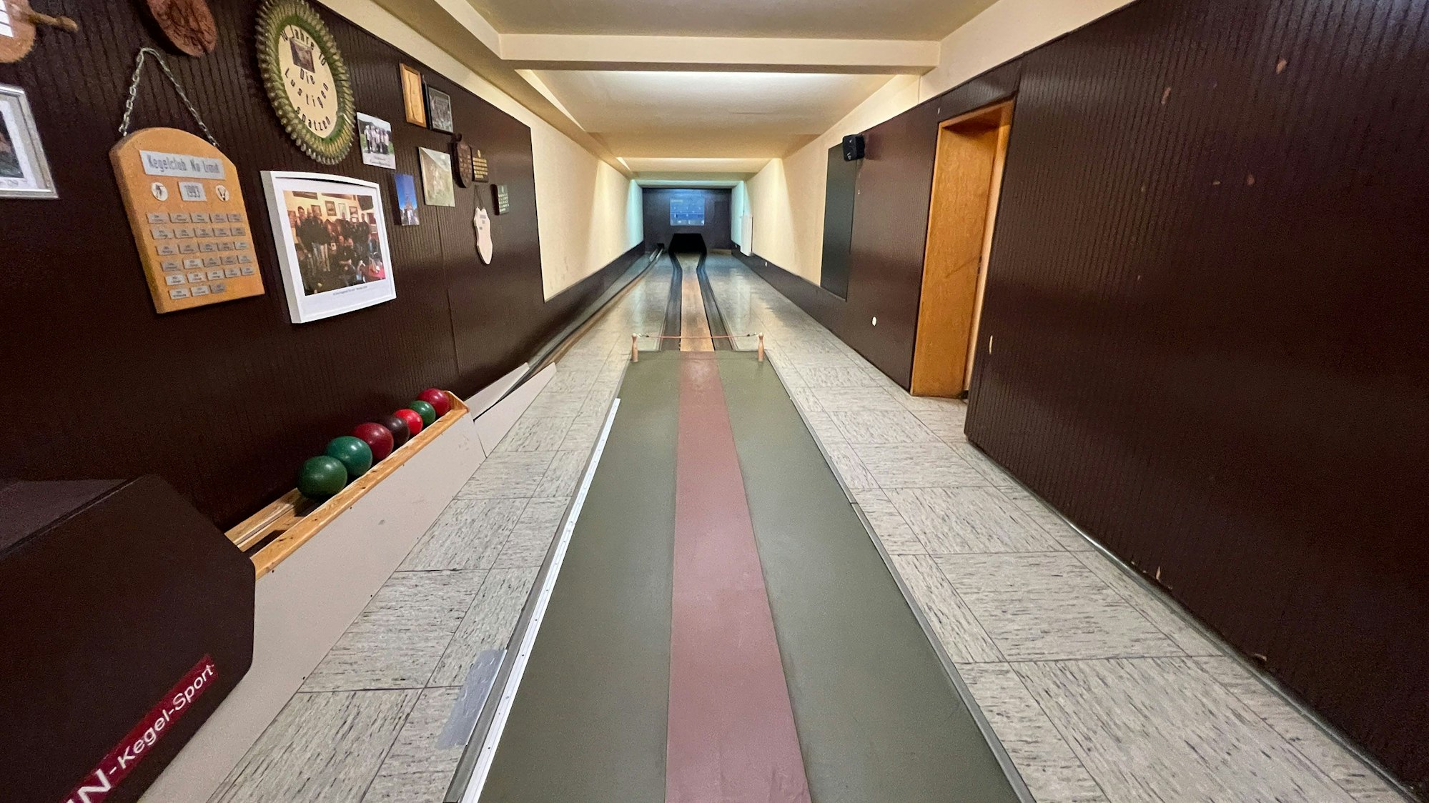 Eine leere Kegelbahn, an den mit dunklem Holz vertäfelten Wänden hängen Bilder und ein Sparkasten.