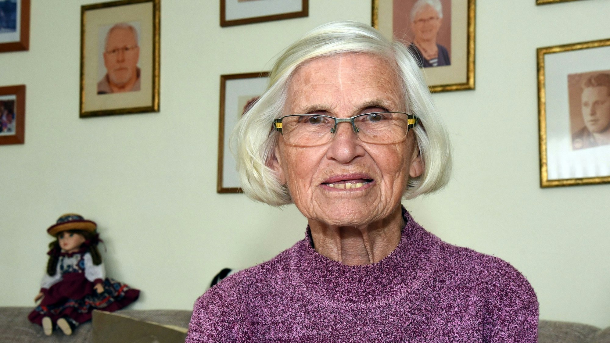 Waltraud Geisen sitzt in ihrem Wohnzimmer auf dem Sofa und schaut in die Kamera, hinter sich an der Wand sind Familienbilder zu sehen.