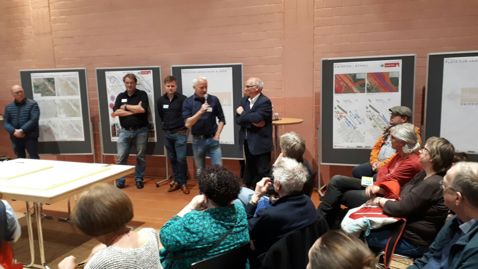 In einem großen Saal stehen Männer mit Namensschildern vor großen Aufstellwänden, im Zuschauerraum sitzen einige Bürger.